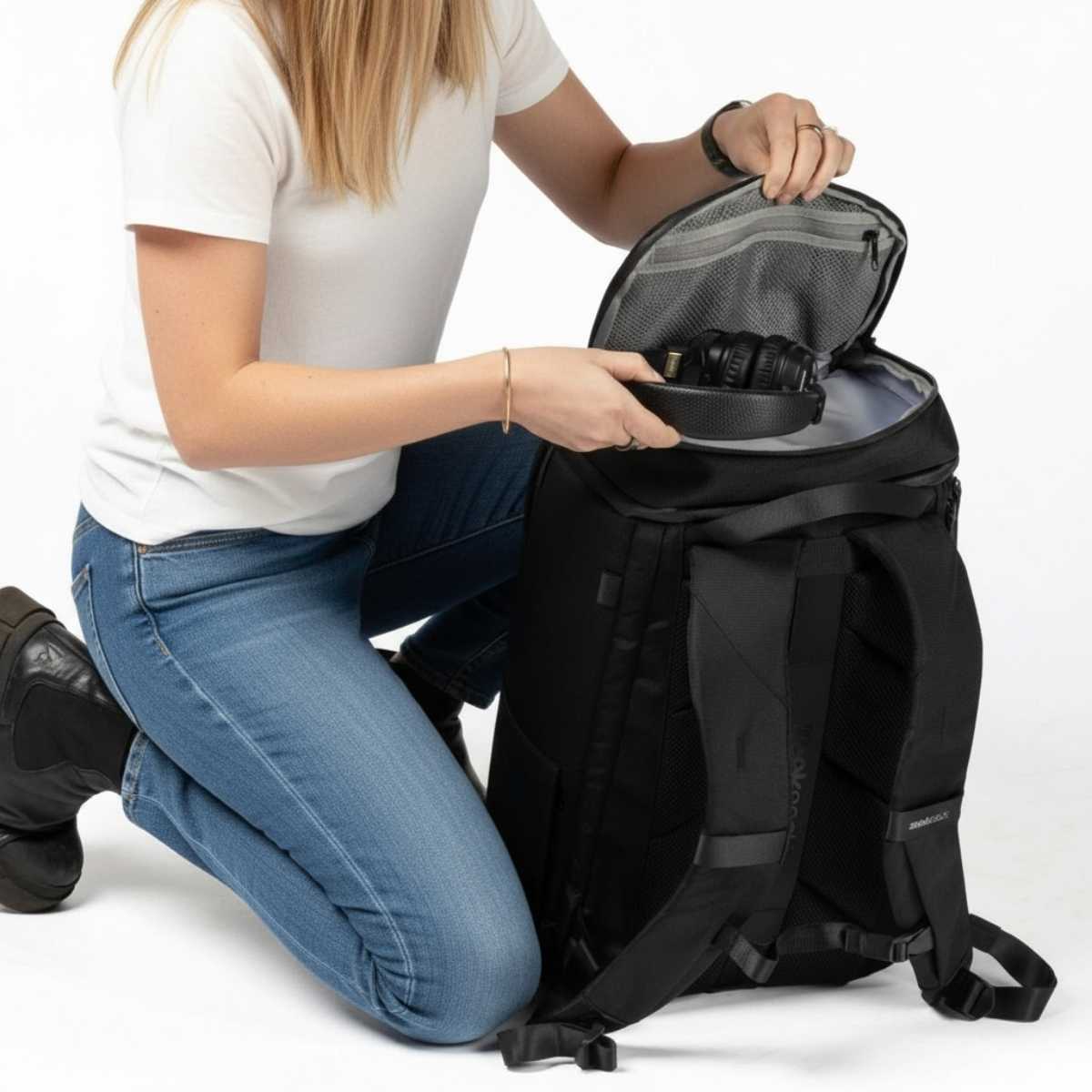 Kvinna i vit t-shirt och jeans sitter på knä och öppnar en svart ryggsäck för att lägga i ett par hörlurar.