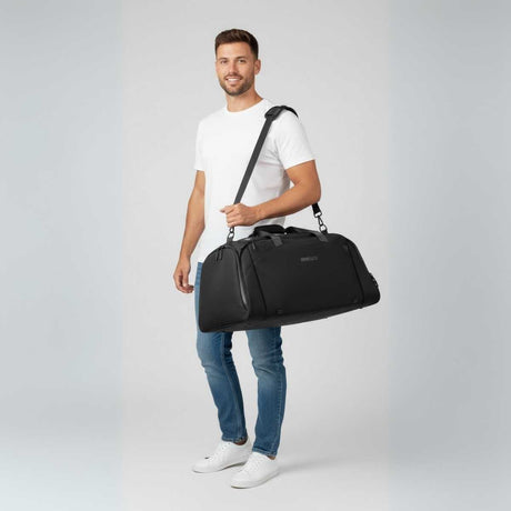 Man i vit t-shirt och jeans bär en svart duffelbag på axeln.