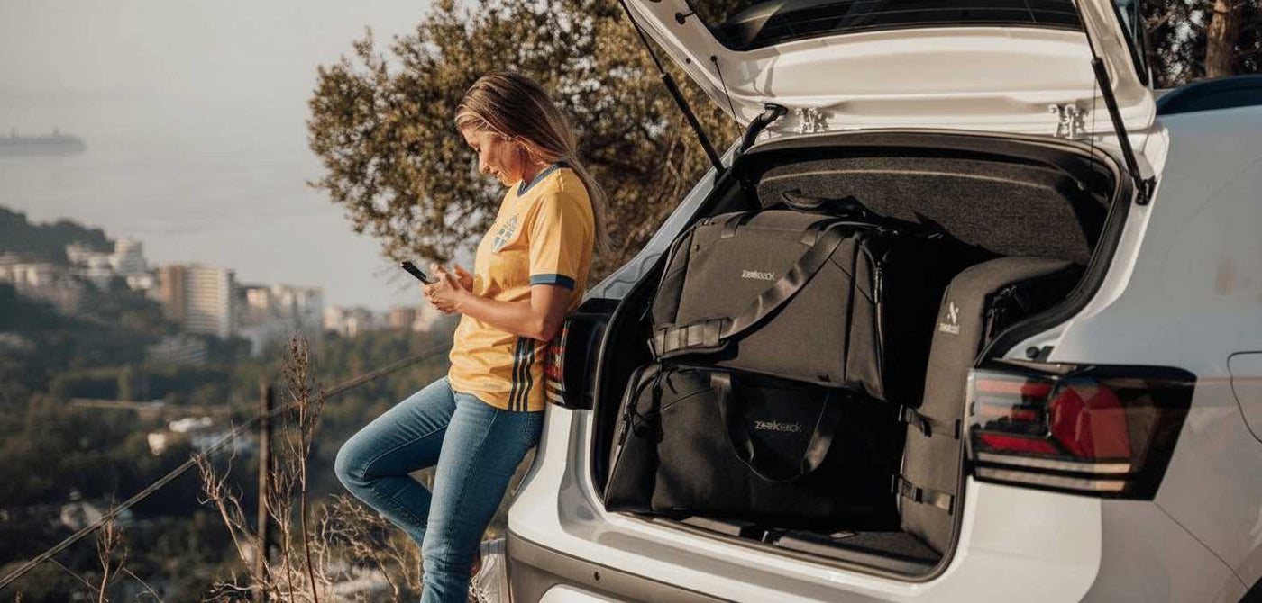 En kvinna i blå jeans, vita sneakers och en gul tröja i svensk landslagsstil lutar sig mot en bil med öppen baklucka. I bagageutrymmet ligger svarta sportväskor och en ryggsäck från Zeeksack. I bakgrunden syns utsikt över stad och hav.
